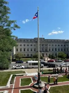 A flag pole in front of a building with people standing around.
