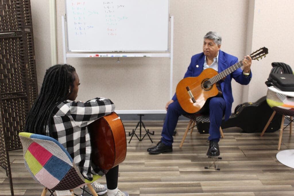 A man in blue jacket playing guitar while sitting on chair.