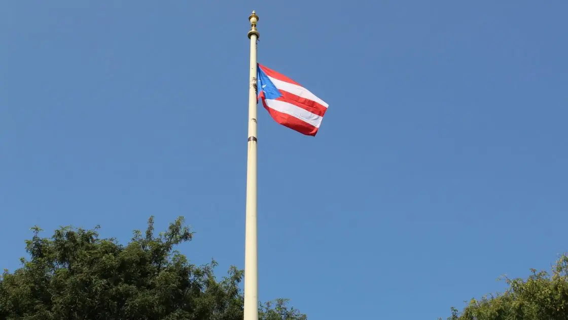 Speaker addressing the crowd at the podium beside the honoree at the Puerto Rican flag raising ceremony in Waterbury