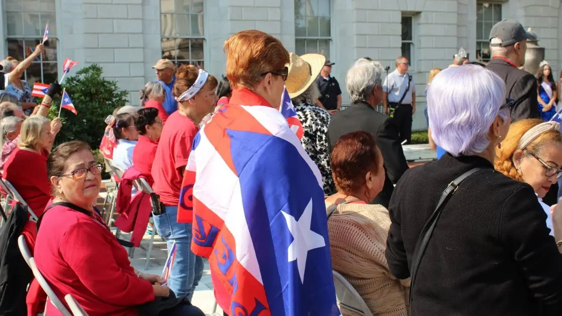 Community members seated at the ceremony, one draped in the Puerto Rican flag, outside Waterbury City Hall